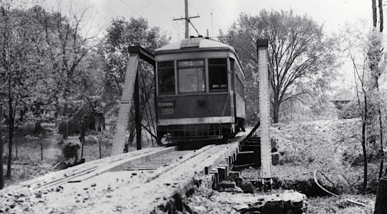 An electric trolley car approaching across a steel truss bridge.