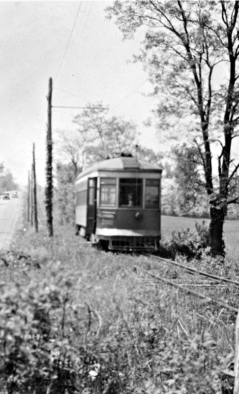 Trolley traveling down the tracks with old automobiles in the distance.